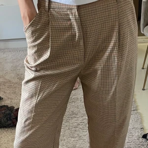 Beige rutiga kostymbyxor wide fit - Snygga beige kostymbyxor med brunt och rött rutmönster. Byxorna har hög midja, vida ben och klassiska pressveck framtill. Mjukt och lätt material med fickor i sidorna, knappt använda, långa på mig som är 175 cm!