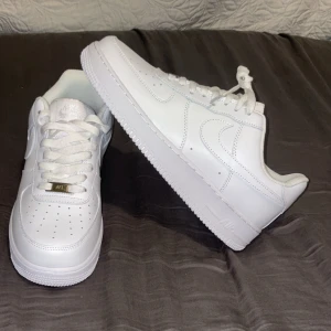 Nike Air Force 1 helvita sneakers - Klassiska Nike Air Force 1 i helvitt skinn med vit sula och snörning. Ikonisk siluett med perforerad tå och metallbricka på snörena. Perfekta för dig som vill ha en clean och tidlös look. Snygga detaljer och bekväm passform.