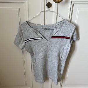 Grå t-shirt Tommy Hilfiger S - Snygg grå t-shirt från Tommy Hilfiger med korta ärmar och v-ringning. T-shirten har svarta, vita och röda ränder över bröstet som ger en sportig vibe. Tillverkad i mjuk bomull, perfekt för en chill och stilren look.