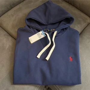 Blå hoodie från Polo Ralph Lauren - Snygg marinblå hoodie från Polo Ralph Lauren med vit snörning och klassisk röd logga broderad på bröstet. Tröjan har huva, känguruficka och ribbade muddar. Perfekt för en chill och stilren look.