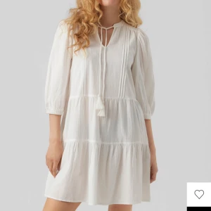 Vit kort klänning med knytband - Superfin vit kortklänning med trekvartsärmar och knytband vid halsen. Klänningen har volangdetaljer och är tillverkad i ett lätt och luftigt bomullstyg, perfekt för varma dagar. Enkel och stilren design som känns både fräsch och trendig.