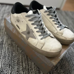 Golden Goose vita sneakers med grå stjärna - Golden Goose sneakers i vitt skinn med grå stjärndetalj på sidan och grå snörning. Skorna har svart hälparti och grå sula. Klassisk låg modell med rund tå och platt sula. Perfekta för en stilren och trendig look.