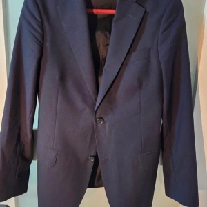 Mörkblå kavaj Oscar Jacobson 48 - Snygg mörkblå kavaj från Oscar Jacobson i klassisk modell. Tillverkad i ulltyg från Vitale Barberis Canonico, med två knappar framtill och flera fickor. Perfekt för dig som vill ha en stilren och tidlös look.