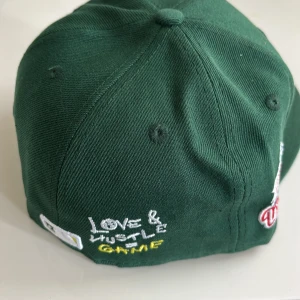 New Era keps 59FIFTY - Snygg grön keps från New Era 59FIFTY med Oakland A's logga broderad framtill och World Series patch på sidan. Kepsen har coola broderade detaljer som 'Love & Hustle = Game' och en liten baseboll. Klassisk rak skärm och Cooperstown Collection-märke på insidan.