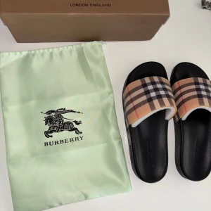 Burberry sandaler - Svarta sandaler från Burberry med bred rem i det ikoniska beige, svarta och röda rutmönstret. Snygg och stilren design med platt sula och rund tå. Tillverkade i Italien och perfekta för en trendig look.