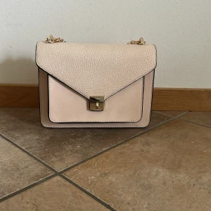 Beige/ljus rosa axelväska med guldkedja - Snygg beige/ ljusrosa axelväska med stilren design och guldfärgad kedja som axelrem. Väskan har ett kuvertformat lock med struktur och en elegant metallspänne framtill. Perfekt storlek för det viktigaste och passar till många outfits.