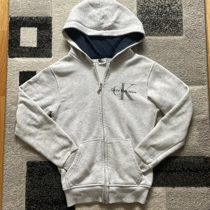 Grå hoodie från Calvin Klein Jeans - Snygg ljusgrå hoodie från Calvin Klein Jeans med dragkedja och stor CK-logga på bröstet. Tröjan har huva med mörkblått foder, två fickor fram och ribbade muddar. Perfekt för chill dagar och enkel att matcha med jeans eller joggers.