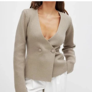 Beige omlott kavaj med knappar - Snygg beige kavaj med omlottknäppning och två stora knappar framtill. Djup v-ringning och långärmad modell i ett mjukt, stickat material. Perfekt för en clean och stilren look. Ifrån Moa matssons kollektion tillsammans med nakd!