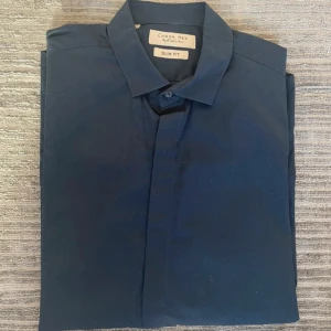 Marinblå slim fit skjorta Chrom Men - En marinblå skjorta i slim fit modell. På skjortan står det att det är storlek XL men jag skulle säga att den motsvarar mer en M storlek.  Fin marinblå färg som inte är urtvättad.   Axelmått 40 cm Längd 73 cm