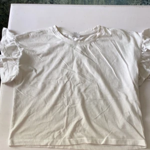 Vit volangtopp från Lindex - Supersöt vit topp från Lindex med volangdetaljer på ärmarna. T-shirten har rund halsringning och är tillverkad i mjuk bomull, perfekt för en chill och stilren look. Passar dig som gillar enkla men ändå unika detaljer.