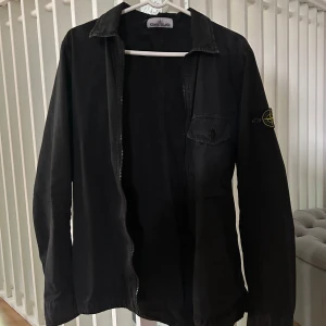 Overshirt Stone Island - Svart overshirt från Stone Island med klassisk krage, dragkedja framtill och bröstficka med knapp. Ikonisk logga på vänster ärm. Perfekt för lager-på-lager och stilren streetwear-vibe. Strl M, mycket bra skick. Självklart äkta!