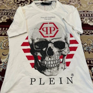 Vit Philipp Plein t-shirt med dödskalle/ byte  - Snygg vit t-shirt från Philipp Plein med stor dödskalle framtill, röda ränder och blingade detaljer. Logga på bröstet och bak på ryggen, samt röd linje längs ryggen. Perfekt för dig som gillar statement-plagg och streetstyle.