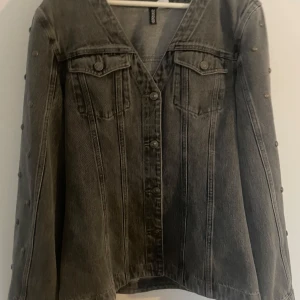 Grå jeansjacka - Snygg grå jeansjacka från hm med coola nitar längs ärmarna och klassiska bröstfickor med knappar. Jackan har v-ringad krage och knäppning framtill. Perfekt för dig som vill ha en edgy och trendig look.