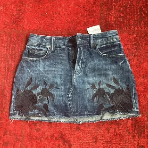 Äkta Guess jeanskjol❤️ - Snygg blå jeanskjol från Guess med rå kant nertill och svarta broderade blommor framtill. Klassisk femficksmodell, dragkedja och knapp i midjan samt Guess-logga på bakfickan. Perfekt för dig som gillar en edgy och trendig stil. Tryck ej köp nu