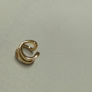 Trendig guldig spiralring - Säljer en snygg ring i guldton med spiralformad design. Ringen har en öppen och modern look med mjuka, svepande linjer som ger en cool och unik känsla. Perfekt accessoar för att lyfta din outfit med lite extra edge.