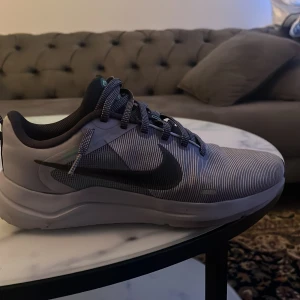 Nike downshifter 12 grå - Snygga Nike sneakers i grått med tunna svarta ränder och svart swoosh-logga på sidan. Skorna har snörning, rund tå och en bekväm platt sula. Perfekta för dig som gillar stilrena och sportiga skor med modern känsla.