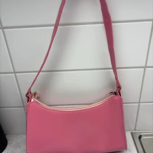 Rosa axelväska i glansigt material - Snygg rosa axelväska med glansig finish och enkel design. Väskan har en ljus dragkedja upptill och ett svart innerfoder. Perfekt storlek för det viktigaste och ger en poppig färg till din outfit.