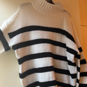 Randig stickad polotröja  - Säljer en stickad polotröja med breda svarta och vita ränder. Tröjan har hög krage och långa ärmar, perfekt för kyliga dagar. Oversized passform för en chill och trendig look.