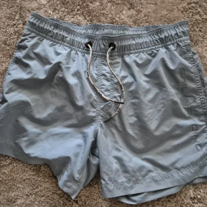 Ljusblå Sail Racing badshorts - Snygga ljusblå badshorts från Sail Racing med elastisk midja och vit snörning framtill. Shortsen har en bakficka med logotyp och är tillverkade i ett snabbtorkande syntetmaterial. Perfekta för stranden eller poolen i sommar!