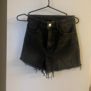 Svarta jeansshorts från H&M Divided - Snygga svarta jeansshorts från H&M Divided med hög midja och råa, fransiga kanter. Klassisk femficksmodell med knapp och dragkedja framtill. Perfekta för sommaren och ger en cool vibe till din outfit.