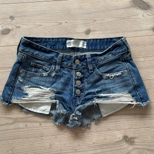 Lågmidjade jeansshorts Abercrombie - Snygga blå jeansshorts från Abercrombie & Fitch med låg midja, slitna detaljer och fransig kant. Knappar framtill och klassiska fickor både fram och bak. Perfekta för sommaren och ger en cool, avslappnad vibe.