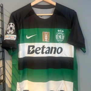 Sporting CP Gyökeres matchtröja Nike - Sporting CP fotbollströja med Gyökeres och nummer 9 på ryggen. Tröjan är randig i svart, grön och vit med klubbmärke, Nike-logga och Betano sponsortryck. Korta ärmar med UEFA Foundation-märke på båda sidor. Perfekt för dig som älskar fotboll och Sporting. Helt ny med tagg en replika