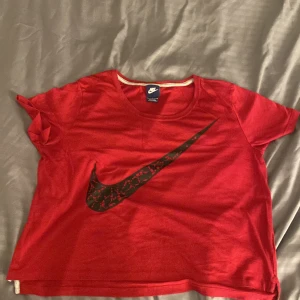 Röd Nike t-shirt med svart logga - Röd t-shirt från Nike med stor svart Swoosh-logga framtill. Klassisk rund halsringning och korta ärmar. Skön passform och tillverkad i mjukt bomullsmaterial. Perfekt för dig som gillar sportig och avslappnad stil.