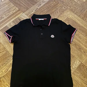 Svart Moncler piké - Svart pikétröja från Moncler med klassisk krage och knäppning framtill. Röd och vit randig detalj på krage och ärmslut samt broderad Moncler-logga på bröstet. Tillverkad i bomull och har en normal passform. Perfekt för en clean och sportig look. Storlek Small. Endast prövad, och insåg direkt att den var för liten. 