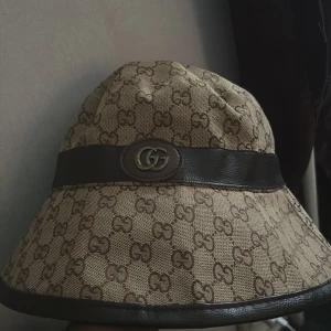 Gucci beige bucket hat med GG-logga - Snygg bucket hat från Gucci i beige canvas med brunt GG-monogram över hela hatten. Hatten har ett brett brunt band i skinnimitation runtom och en GG-logga framtill. Perfekt accessoar för att lyfta din outfit med lyxig känsla.