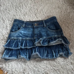 Blå volang jeanskjol med slitningar - Supersöt kort jeanskjol i blå denim med två lager volanger och råa kanter. Kjolen har klassiska fickor fram och bak, bälteshällor och knäppning framtill. Sitter jättefint. Är storlek 158 men passar större! 
