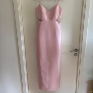 Ljusrosa långklänning med cut-outs - Supersnygg ljusrosa långklänning med smala axelband, v-ringning och trendiga cut-outs i midjan. Klänningen har en slits baktill och är gjord i ett glansigt satinliknande material som ger en lyxig känsla. Perfekt för dig som vill sticka ut med en elegant och modern look. Klänningen är använd 1ggr  i några timmar.