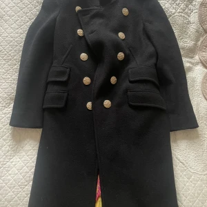 Svart skepparkavaj från FEMME by Michele R. - Stilren svart skepparkavaj med dubbelknäppning i guld, dekorativa fickor och kedjedetalj i ryggen. Jackan har lång ärm och är fodrad med blommigt tyg. Perfekt för dig som vill ha en klassisk men edgy look.