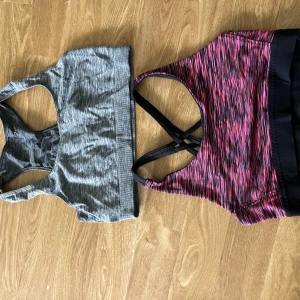 2 sportiga linnetoppar i grått och rosa - Två sportiga linnetoppar, en i melerad grå och en i rosa med svarta och lila inslag. Båda har brottarrygg, ärmlös design och bred resår nedtill. Perfekta för träning och aktiva dagar. Den rosa har snygga korsade band i ryggen för extra stilpoäng.