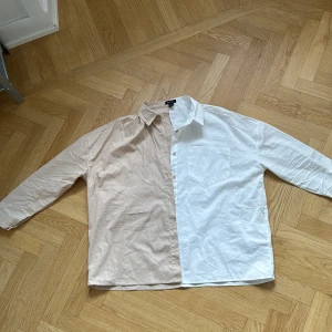 Beige och vit skjorta från Boohoo - Säljer en snygg tvåfärgad skjorta från Boohoo i beige och vitt. Skjortan har klassisk krage, lång ärm och bröstficka på den vita sidan. Tillverkad i mjuk bomull och har knappar framtill. Perfekt för dig som gillar en unik och trendig look.