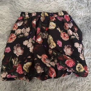 Blommig kortkjol från Creamie - Supersöt kortkjol från Creamie med svart botten och färgglatt blommönster i rosa, rött, gult och grönt. Kjolen har en luftig och voluminös form med resår i midjan. Perfekt för dig som gillar romantiska vibes och vill sticka ut lite extra.