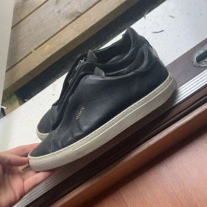 Svarta sneakers från Axel Arigato - Stilrena svarta sneakers från Axel Arigato i skinn med vit platt sula och dragkedja framtill istället för klassisk snörning. Minimalistisk design med diskret guldtext på sidan. Perfekta för dig som gillar clean och modern look.