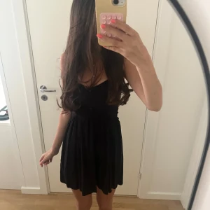 Svart kort klänning med knappar - Supersöt svart kortklänning med knappar framtill och smickrande v-ringning. Klänningen har en lätt utställd kjol och smala axelband, perfekt för varma dagar. Enkel och stilren design