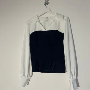 Vit och marinblå korsettblus - Snygg tvåfärgad skjort/blus med vit skjortkrage och långa ärmar, kombinerat med en marinblå korsettliknande midja. Ballongärmar med breda manschetter och markerad söm framtill ger en cool och trendig look. Perfekt för dig som gillar att klä dig elegant och proffsigt! Den är i mycket bra skick aldrig använd då den är för stor för mig!