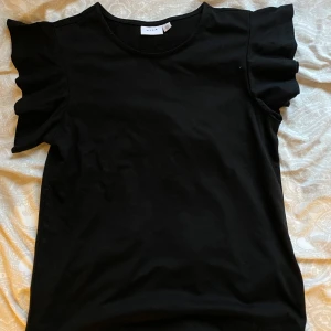 Svart topp med volangärmar från Vila - Snygg svart topp från Vila med korta volangärmar som ger en extra touch till outfiten. Enkel rund halsringning och mjukt bomullsmaterial. Perfekt för dig som gillar stilrena plagg med en twist.