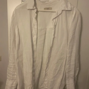 Vit linneskjorta från Gina Tricot - Klassisk vit skjorta från Gina Tricot i svalt och luftigt linne. Skjortan har lång ärm, krage och knäppning framtill. Perfekt för en clean och stilren look, lätt att matcha med jeans eller kjol.