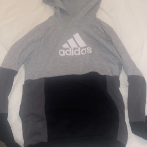 Grå och svart hoodie från Adidas - Snygg hoodie från Adidas i grått och svart med stor vit logga på bröstet. Tröjan har huva och långa ärmar, samt paneler i olika grå och svarta nyanser. Perfekt för en sportig och chill stil.