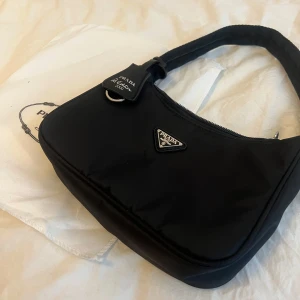 Svart axelväska  - Säljer en svart axelväska  med klassisk trekantig logga i metall framtill. Väskan är i mjukt nylonmaterial och har en enkel, stilren design med dragkedja upptill och bred axelrem. Perfekt för att lyfta vilken outfit som helst. Finns en liten repa men syns knappt. Dm för mer bilder 