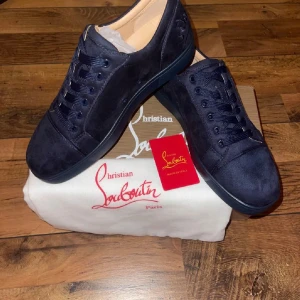 Mörkblå Louboutins  - Snygga mörkblå sneakers från Christian Louboutin i mocka med klassisk röd sula och diskret logga på hälen. Skorna har rund tå, platt sula och matchande mörkblå snörning. Perfekta för dig som vill sticka ut med exklusiv stil.