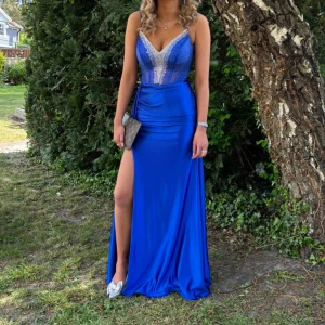 Blå långklänning med slits och glitter - Supersnygg blå långklänning med djup v-ringning och glittriga detaljer på livet. Klänningen har en hög slits på ena sidan och smala axelband. Materialet är glansigt och följsamt, perfekt för bal eller fest. Sitter figurnära upptill och faller snyggt nedtill.