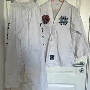 Taekwondo dräkt Budo-Nord storlek 2/150 - Taekwondo dräkt från Budo-Nord i vitt med svarta tryck och broderade märken på jackan. Setet innehåller jacka, byxor samt vitt och gult bälte. Materialet är 55% bomull och 45% polyester. Perfekt för dig som tränar taekwondo och vill ha en klassisk look.