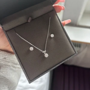 Silverset med halsband & örhängen Albrekts Guld - Snyggt smyckeset från Albrekts Guld i äkta silver med halsband och matchande örhängen. Båda har glittrande stenar av kubisk zirkonia i blomformad design. Kedjan är tunn och elegant, perfekt för att ge en extra touch till din look. Asken följs med.