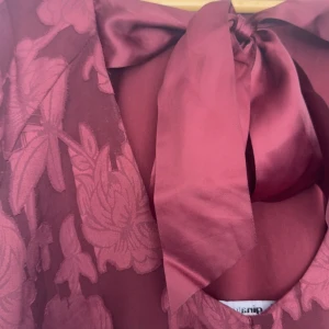 Vinröd blommig klänning med rosett - Vinröd långärmad klänning från Gina Tricot med blommigt mönster i tyget och stor rosett i ryggen. Klänningen har U-ringning och är gjord i ett lätt, transparent material med ett innerfoder. Perfekt för dig som vill sticka ut med en romantisk vibe.