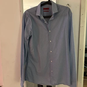 Blå extra slim fit skjorta Hugo Boss - Snygg blå skjorta från Hugo Boss i extra slim fit-modell. Skjortan har ett diskret mönster, klassisk krage och vita knappar framtill. Perfekt för dig som gillar en tajtare passform och stilren look.