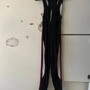 Svart sportig jumpsuit med dragkedja - Sportig jumpsuit i svart med vita och vinröda paneler längs sidorna. Modellen är ärmlös med djupa ärmhål och har en dragkedja framtill. Perfekt för träning eller danspass. Materialet är stretchigt och följsamt för maximal rörelsefrihet.