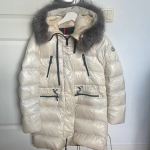 Vit Moncler dunjacka med pälskant, strl M - Snygg vit dunjacka från Moncler med äkta pälskant på huvan och svarta detaljer. Jackan har flera dragkedjefickor, justerbar midja med snörning och quiltad design. Perfekt för kalla dagar när du vill vara både varm och stilren. Storlek 2 i moncler vilket motsvarar M.